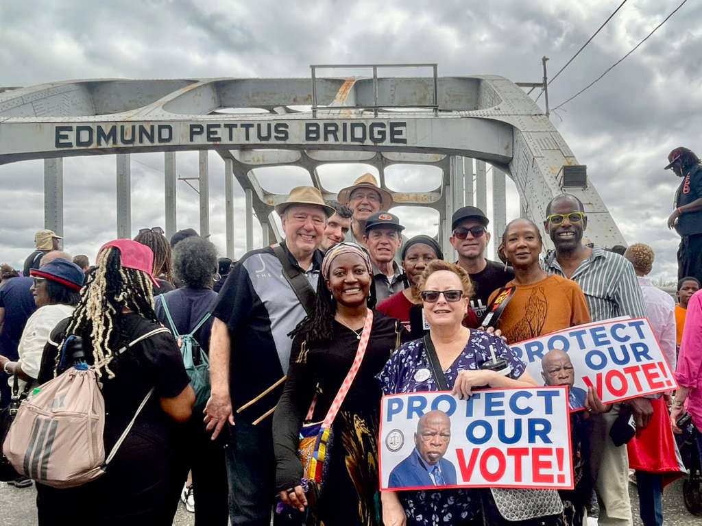 The Words cast stands on the Edmund Pettus Bridge, holding signs that read 'PROTECT OUR VOTE!' Some signs also feature an image of John Lewis.